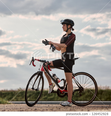 Fit woman in cycling gear standing beside red and black road bike, calmly putting on gloves under cloudy sky, preparing before training. Fit woman in cycling gear standing beside red and black road bike, calmly putting on gloves under cloudy sky, preparing before training. 126452928