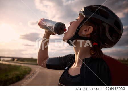 Close-up photo of female cyclist pouring water from bottle into mouth, sunlight behind and road curving in distance, drops splashing in midair. 126452930