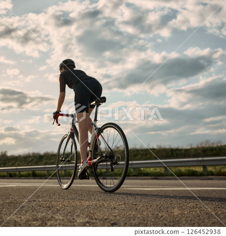 Rear view photo of woman cycling on empty road surrounded by soft clouds and flat landscape, showing solo ride atmosphere and determination in posture. Rear view photo of woman cycling on empty road surrounded by soft clouds and flat landscape, showing solo ride atmosphere and determination in posture. 126452938
