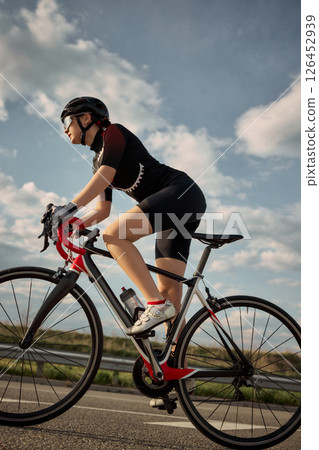Side view photo of cyclist riding road bike under bright sky, pushing forward on clean highway, captured in golden light with focus on pedaling motion. Side view photo of cyclist riding road bike under bright sky, pushing forward on clean highway, captured in golden light with focus on pedaling motion. 126452939