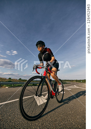 Low angle view photo of female cyclist riding on curvy road, hands on lower handlebars, showing aggressive racing posture and focus in eyes. Low angle view photo of female cyclist riding on curvy road, hands on lower handlebars, showing aggressive racing posture and focus in eyes. 126452943