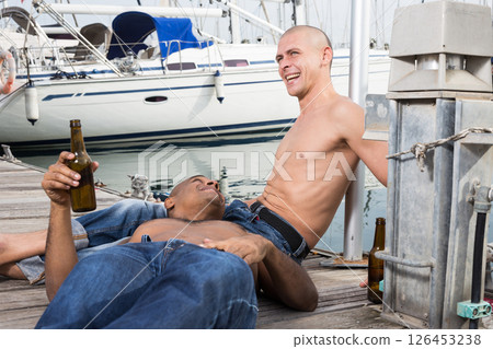 Romantic couple of young men sitting and relaxing together on the pavement in the port Romantic couple of young men sitting and relaxing together on the pavement in the port 126453238