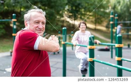 Staying after retirement. Happy joyful mature retired sportsman wearing sportswear doing side stretching exercises with arm over his head, exercising outside in city park 126453269