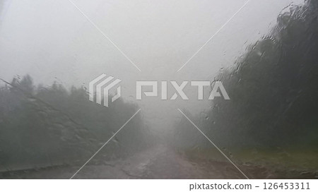 Heavy rain blurs view through car windshield, obscuring road ahead and creating misty atmosphere. Water droplets create textured effect on window, making visibility challenging Heavy rain blurs view through car windshield, obscuring road ahead and creating misty atmosphere. Water droplets create textured effect on window, making visibility challenging 126453311
