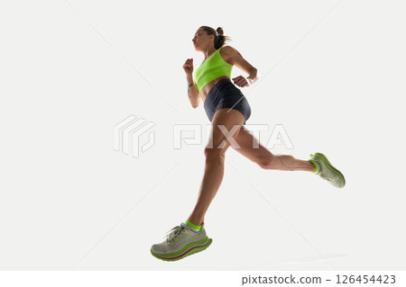 Low angle dynamic image of sportive young woman in green top and black shorts running on white background 126454423