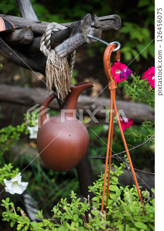 A rustic clay jug hangs from a wooden structure with a rope, surrounded by green plants and vibrant flowers. Concept of rural charm and nature 126456075
