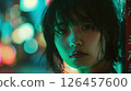 A young woman gazes thoughtfully at the camera under colorful neon lights in a bustling urban setting at night 126457600