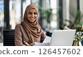 Smiling woman in a hijab working on a laptop in a modern office environment during daytime 126457602