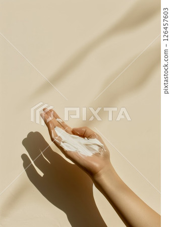 A hand holding a creamy skincare product against a soft beige background with gentle shadows in natural light 126457603
