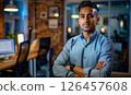 Young professional in a modern office setting confidently poses with arms crossed in a tech-driven workspace during the evening 126457608