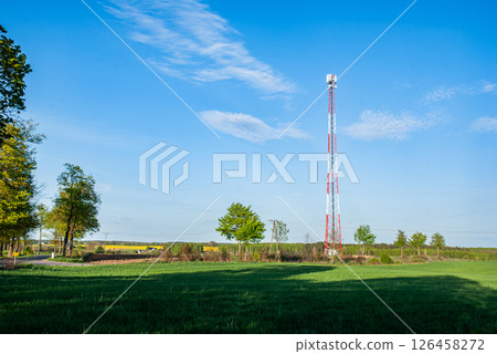 Tall communication tower in lush green field with clear blue sky and scattered clouds amidst rural landscape countryside setting Tall communication tower in lush green field with clear blue sky and scattered clouds amidst rural landscape countryside setting 126458272