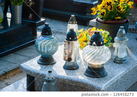 Peaceful cemetery scene with glass lanterns and vibrant flowers on a granite tombstone illuminated by sunlight Peaceful cemetery scene with glass lanterns and vibrant flowers on a granite tombstone illuminated by sunlight 126458273
