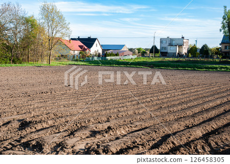 Expansive tilled field stretching towards distant houses under clear blue sky signaling spring's arrival in rural countryside 126458305