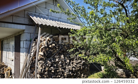 Pile of chopped firewood neatly stacked under a shed beside a country house, with leafy tree in foreground. Concept of self-sufficiency and rural lifestyle 126459332