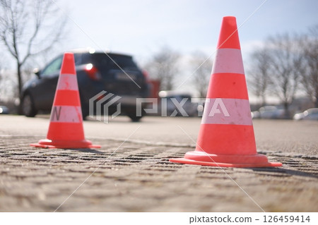 Two orange and white traffic cones are on the ground in front of a car Two orange and white traffic cones are on the ground in front of a car 126459414