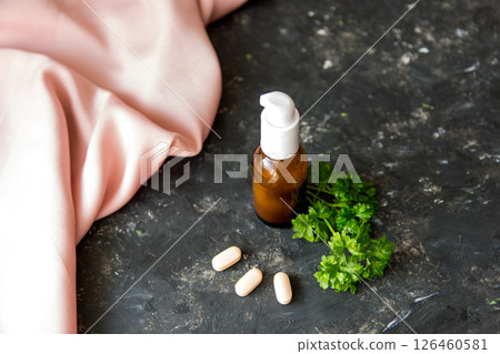 Brown glass bottle with essential oil or creme, vitaminen and healing green herbs on the rough texture background with pink silk fabric. 126460581