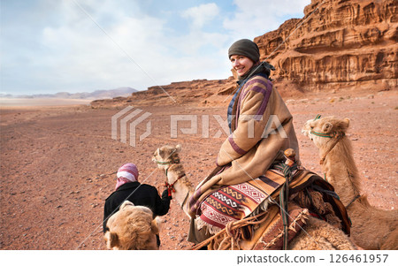 Young woman riding camel in Wadi Rum desert, looking back over her shoulder, smiling. It's quite cold so she is wearing traditional Bedouin coat - bisht - and head scarf, mountains far background 126461957
