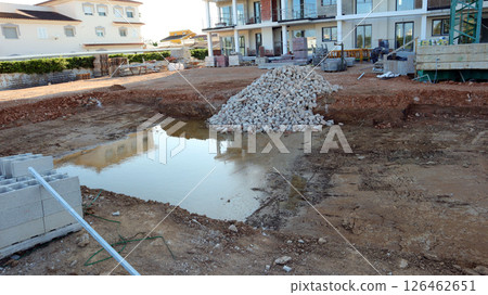 Construction workers are digging a swimming pool near a nearly complete house. The area shows a pit filled with water and a pile of stones, setting the stage for further development. 126462651