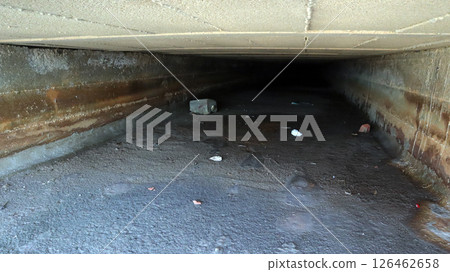 An old drainage channel sits unused, filled with debris and stagnant water. The structure shows wear, with a dark tunnel leading away into obscurity, indicating neglect over time. 126462658