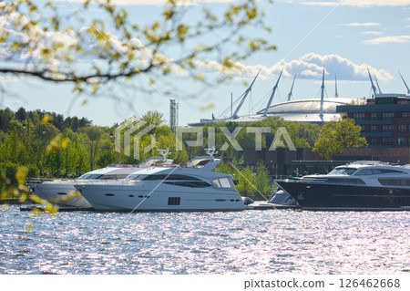 View of the mega yachts standing at the pier through the branches of trees on a clear sunny day, residential buildings and a stadium in the background, Russia, St.Petersburg 126462668