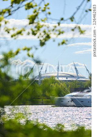 View of the mega yachts standing at the pier through the branches of trees on a clear sunny day, residential buildings and a stadium in the background, Russia, St.Petersburg View of the mega yachts standing at the pier through the branches of trees on a clear sunny day, residential buildings and a stadium in the background, Russia, St.Petersburg 126462683