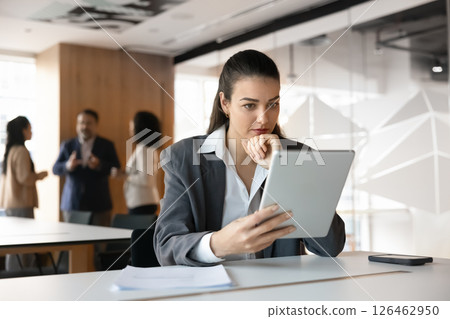 Businesswoman in suit seated at desk looks at tablet screen Businesswoman in suit seated at desk looks at tablet screen 126462950