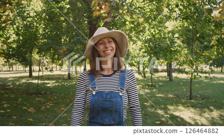 A woman wearing a striped long-sleeve shirt and denim overalls smiles in a vibrant park filled with green trees and autumn-colored leaves on a sunny day. 126464882