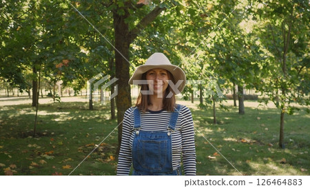 A woman wears a wide-brimmed hat and striped shirt, standing in a vibrant park filled with lush trees and fallen leaves on a sunny autumn day. A woman wears a wide-brimmed hat and striped shirt, standing in a vibrant park filled with lush trees and fallen leaves on a sunny autumn day. 126464883