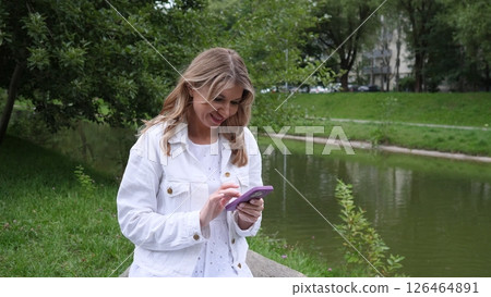A woman dressed in a light jacket smiles as she interacts with her smartphone, seated near a serene river in a green park during daytime. 126464891