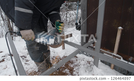 A person operates an angle grinder on a metal frame in a snowy outdoor setting, focused on completing a winter construction task amidst a cold environment. A person operates an angle grinder on a metal frame in a snowy outdoor setting, focused on completing a winter construction task amidst a cold environment. 126464897