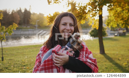 Smiling woman holds a small dog wrapped in a blanket as they share a warm moment in a park surrounded by colorful autumn leaves and a calm lake. Smiling woman holds a small dog wrapped in a blanket as they share a warm moment in a park surrounded by colorful autumn leaves and a calm lake. 126464899