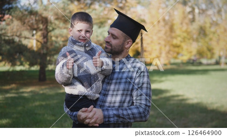 A father celebrates his graduation in a park, holding his young son who is excitedly giving thumbs up. The background features vibrant autumn foliage. A father celebrates his graduation in a park, holding his young son who is excitedly giving thumbs up. The background features vibrant autumn foliage. 126464900