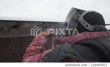 A worker in winter gear carefully uses a torch to melt snow off a roof, surrounded by a cold, snowy landscape in a remote location during wintertime. 126464921