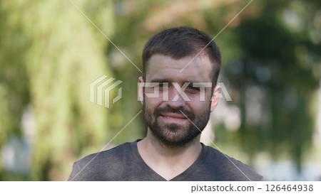 A man with a beard stands outdoors, smiling warmly. Sunlight filters through trees, creating a serene ambiance, perfect for casual socializing or portrait sessions. 126464938