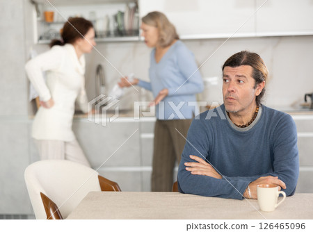 Old woman is quarreling with woman while sad man is sitting with them in kitchen 126465096