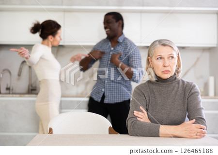 Upset mature woman sitting at table while married couple quarreling in background Upset mature woman sitting at table while married couple quarreling in background 126465188