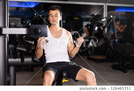 Slender athletic man work out muscles of arms and shoulders on simulator in gym of fitness club 126465351