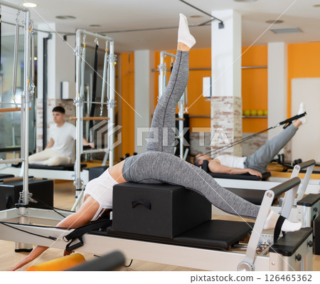Girl works out in gym and performs set of exercises to stretch leg muscles with help of reformer Girl works out in gym and performs set of exercises to stretch leg muscles with help of reformer 126465362