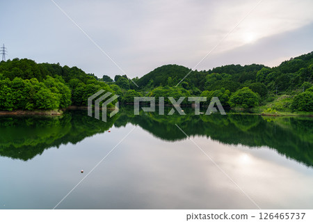 The sun peeking through the thin clouds and the sky reflected on the mirror-like lake surface at Nunome Dam in Nara City, Nara Prefecture➅ 126465737
