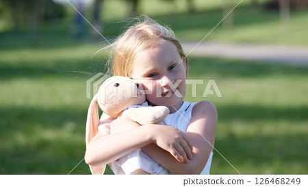 Young girl with long blonde hair embraces a soft bunny toy, showcasing joy in a bright park setting filled with grass and trees during a sunny day. Young girl with long blonde hair embraces a soft bunny toy, showcasing joy in a bright park setting filled with grass and trees during a sunny day. 126468249