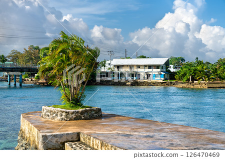 Coastal view from Long Island Park in Koror, Palau, showing a small group of palm trees on a stone platform by the water with buildings and a bridge across the channel in the background under a partly Coastal view from Long Island Park in Koror, Palau, showing a small group of palm trees on a stone platform by the water with buildings and a bridge across the channel in the background under a partly 126470469
