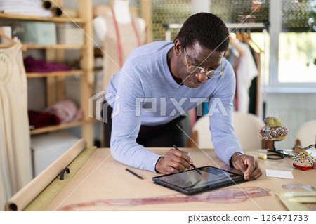 Male tailor draws sketch of future clothing on an electronic tablet. Working with modern gadgets in sewing workshop Male tailor draws sketch of future clothing on an electronic tablet. Working with modern gadgets in sewing workshop 126471733