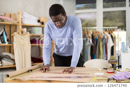 Male tailor draws sketch of future clothing on paper using patterns in sewing workshop Male tailor draws sketch of future clothing on paper using patterns in sewing workshop 126471740