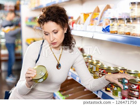 In Russian goods store, woman choose canned sprats in oil 126471750
