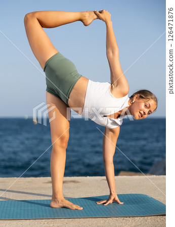 On windy day young slender woman does yoga near sea doing Natarajasana 126471876
