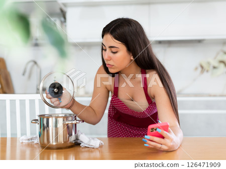 Young woman holding mobile phone opening lid of the pan in the kitchen 126471909