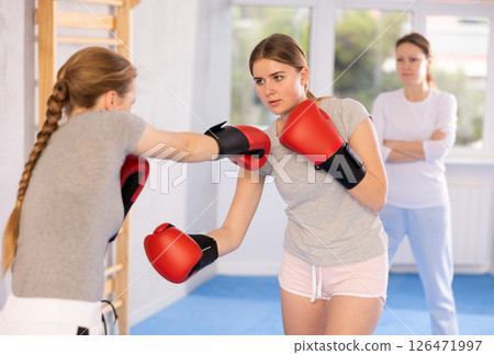 Two teenage girls in sportswear are engaged in boxing sparring in the gym under guidance of trainer in gym 126471997