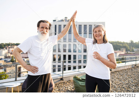 Elderly man and woman celebrating fitness exercise with a high five on rooftop. Outdoors setting with urban skyline in background. Concept of health, happiness, and active lifestyle in senior age. Elderly man and woman celebrating fitness exercise with a high five on rooftop. Outdoors setting with urban skyline in background. Concept of health, happiness, and active lifestyle in senior age. 126472012