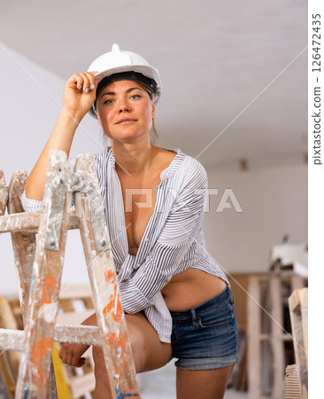 Seductive woman in hardhat, denim shorts and shirt tied on naked tummy standing in apartment during overhauls 126472435