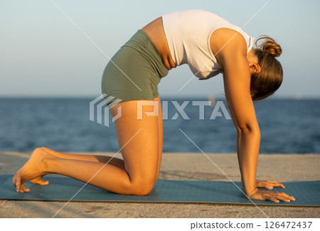 On windy day young slender woman does yoga near sea doing Marjariasana, cat pose 126472437